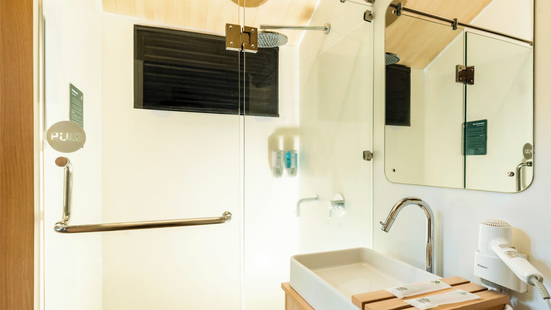 A modern bathroom with a glass shower enclosure, a pull bar, a sink with a mirror, and a hairdryer.