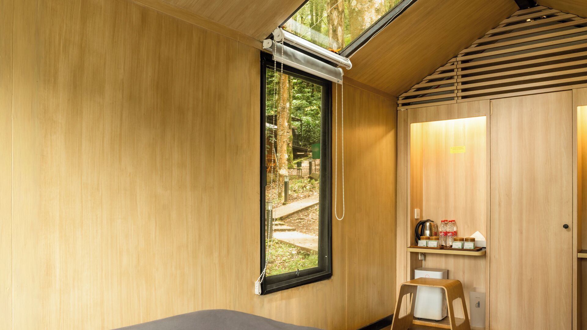Una habitación de madera con tragaluz, una cama, una mesita y una estantería con frascos.