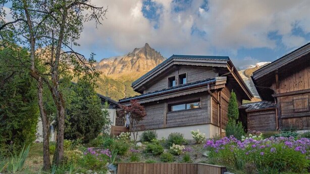 A rustic wooden chalet with a stone pathway leading to the entrance, surrounded by lush greenery and a mountain backdrop.