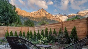 A wooden deck with two black chairs and a wooden fence with coniferous trees.