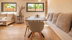 A modern living room with a wooden dining table, a sofa, and a bench. There are two windows providing views of greenery outside.