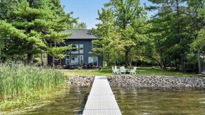 A house with a dock extending into a pond, surrounded by trees and a grassy area with outdoor furniture. - New lake Retreat 1.5 hours from Twin cities in the heart of WI cabin country! (Frederic)