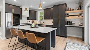 A modern kitchen with dark cabinets, a central island with bar stools, pendant lighting, and a coffee station. - New lake Retreat 1.5 hours from Twin cities in the heart of WI cabin country! (Frederic)