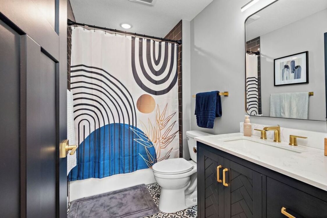 A modern bathroom with a shower curtain featuring abstract patterns, a white toilet, a vanity with a mirror, and a towel rack.