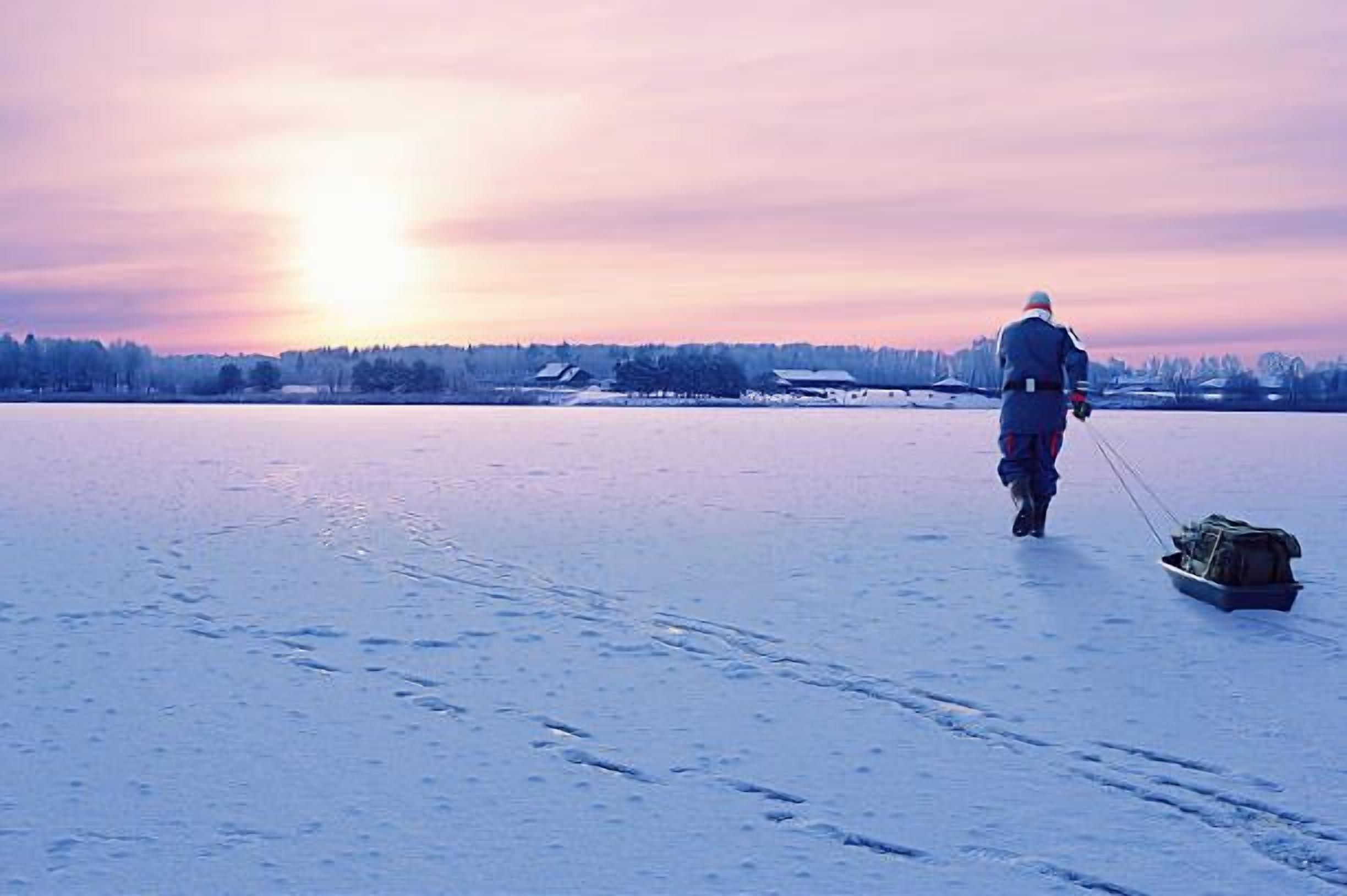 A person walking on a frozen lake with a sled, surrounded by snow-covered trees and buildings in the distance.