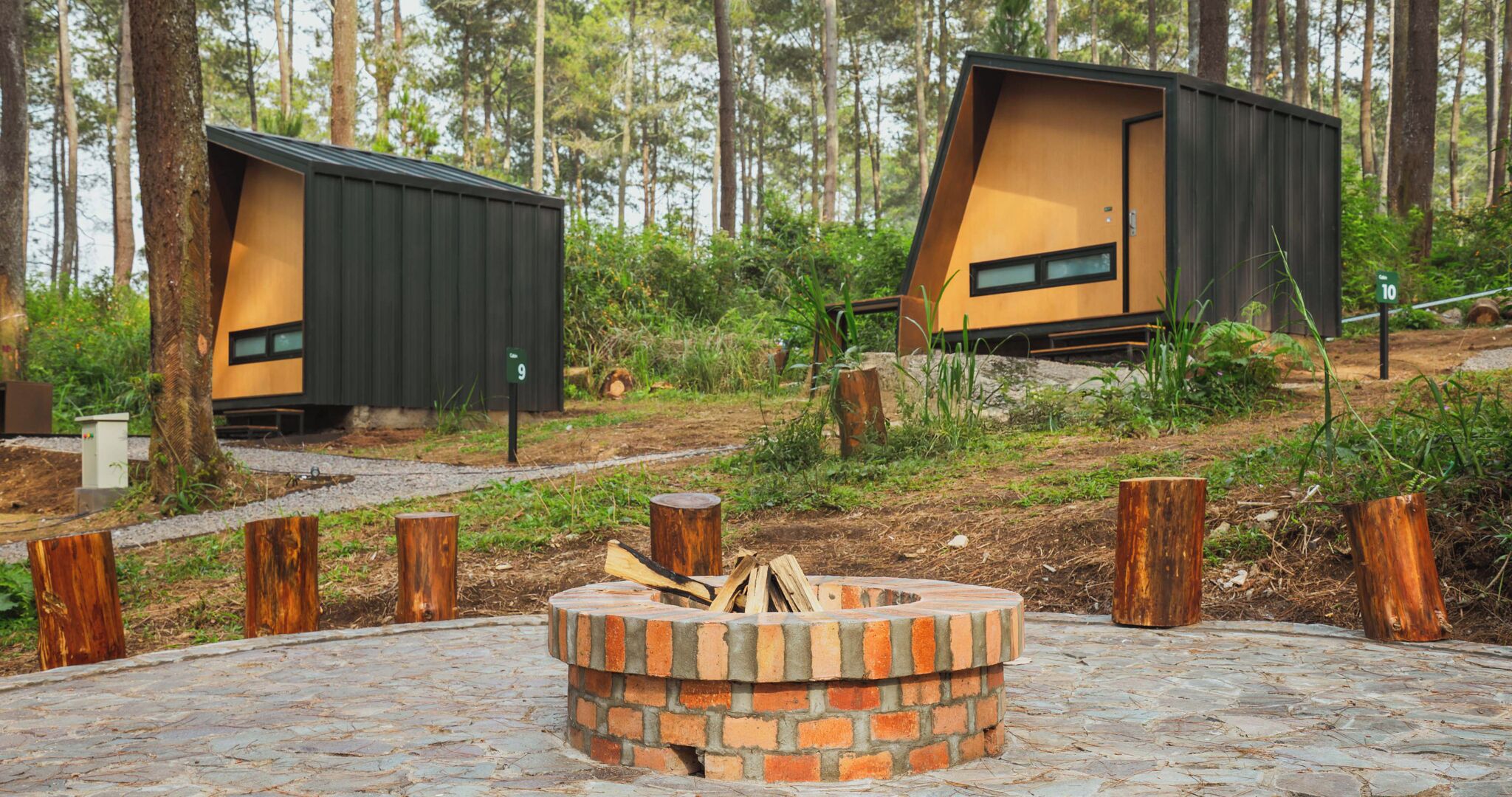 Two modern cabins with sloped roofs, surrounded by a forest. A brick fire pit is in the foreground.