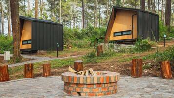 Two modern cabins with sloped roofs, surrounded by a forest. A brick fire pit is in the foreground.