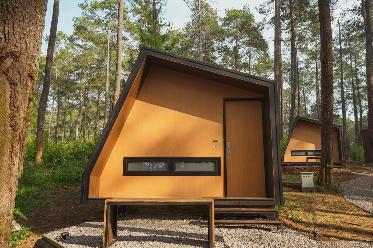 Uma cabana moderna em formato triangular, com uma janela grande e uma porta, localizada em uma área arborizada com chão de cascalho.