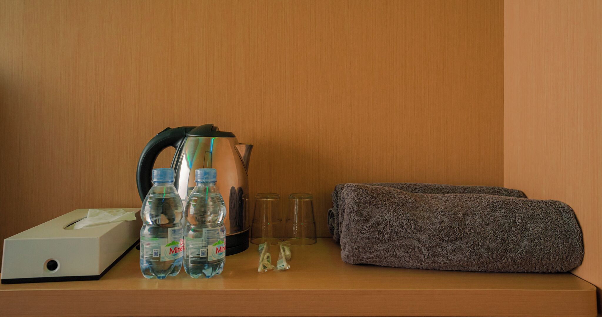A hotel room with a wooden nightstand holding a tissue box, two water bottles, a kettle, two glasses, and a folded towel.