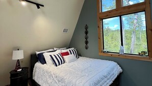 Een slaapkamer met een schuine kap, een bed met witte beddengoed en gestreepte kussens, een nachtkastje met een lamp en een raam met uitzicht op bomen.