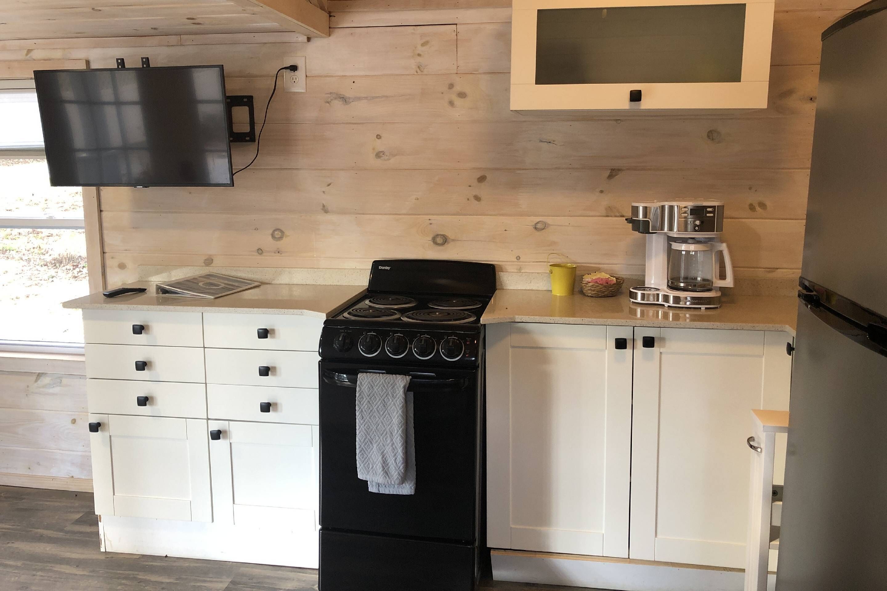 A kitchen with a black stove, white cabinets, a coffee maker, and a flat-screen TV mounted on the wall.