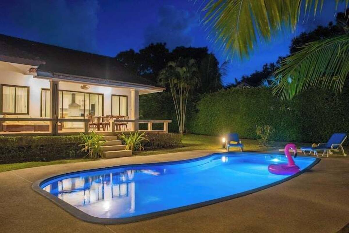 A swimming pool with a pink flamingo float, a covered patio with outdoor furniture, and a house with large windows and a well-lit exterior.