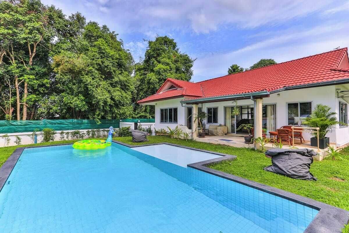 A house with a swimming pool, surrounded by greenery and a red-roofed building.