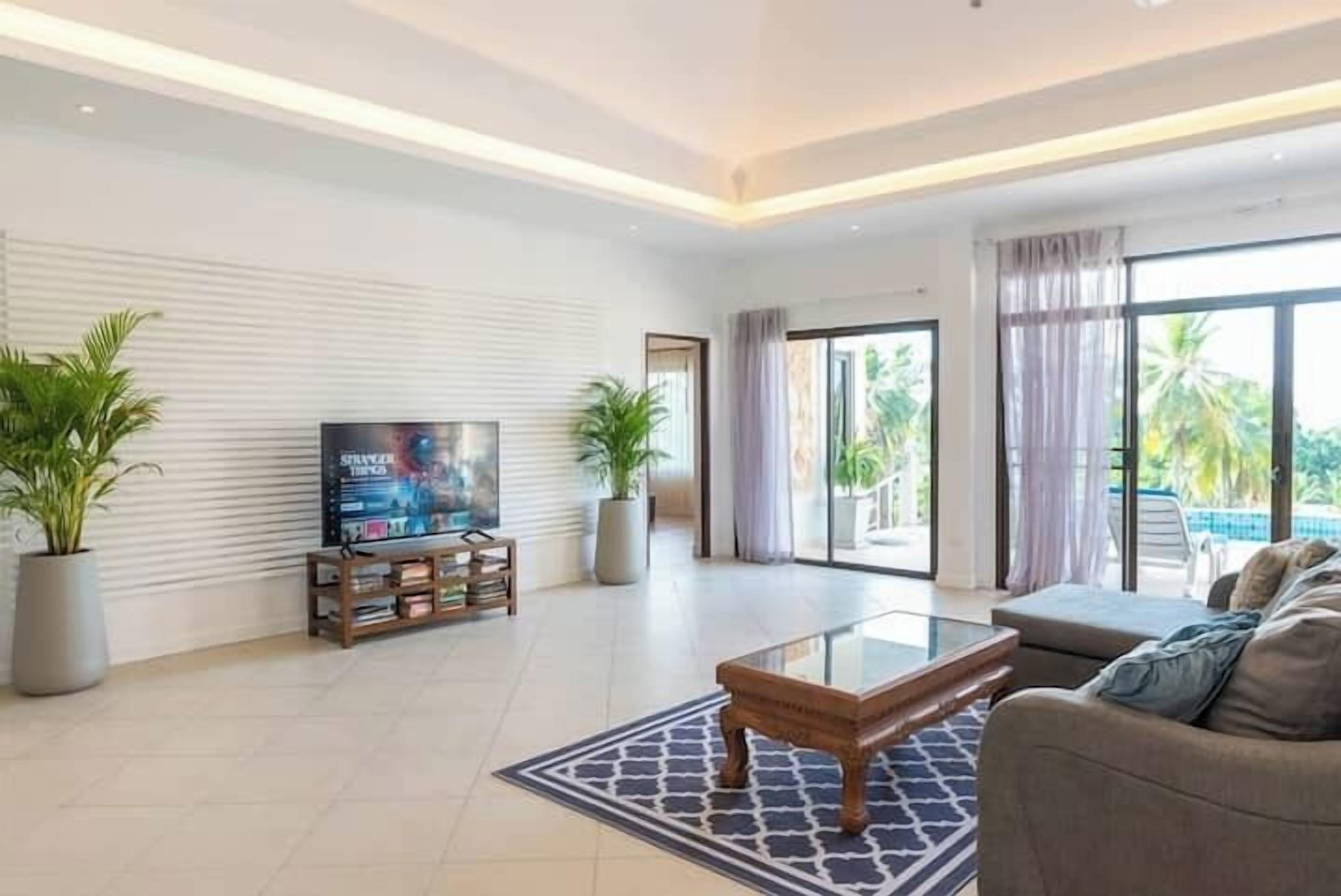 A modern living room with a flat-screen TV, a glass coffee table, and a sofa. There are potted plants and a large window with a view of palm trees.