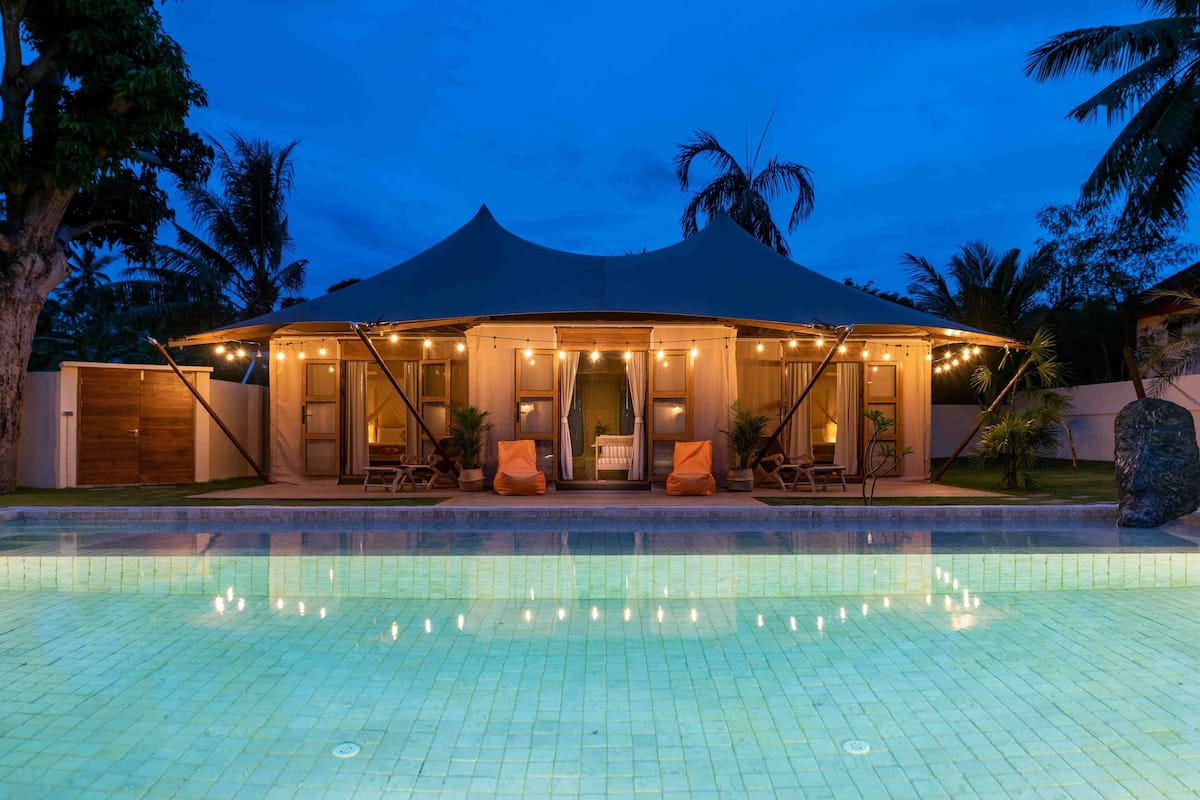 A poolside area with a covered seating area, a pool, and palm trees in the background.