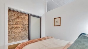 Ein Schlafzimmer mit Backsteinwand, einer Schiebetür, einem Bett mit beiger und grüner Decke sowie einem gerahmten Bild an der Wand.
