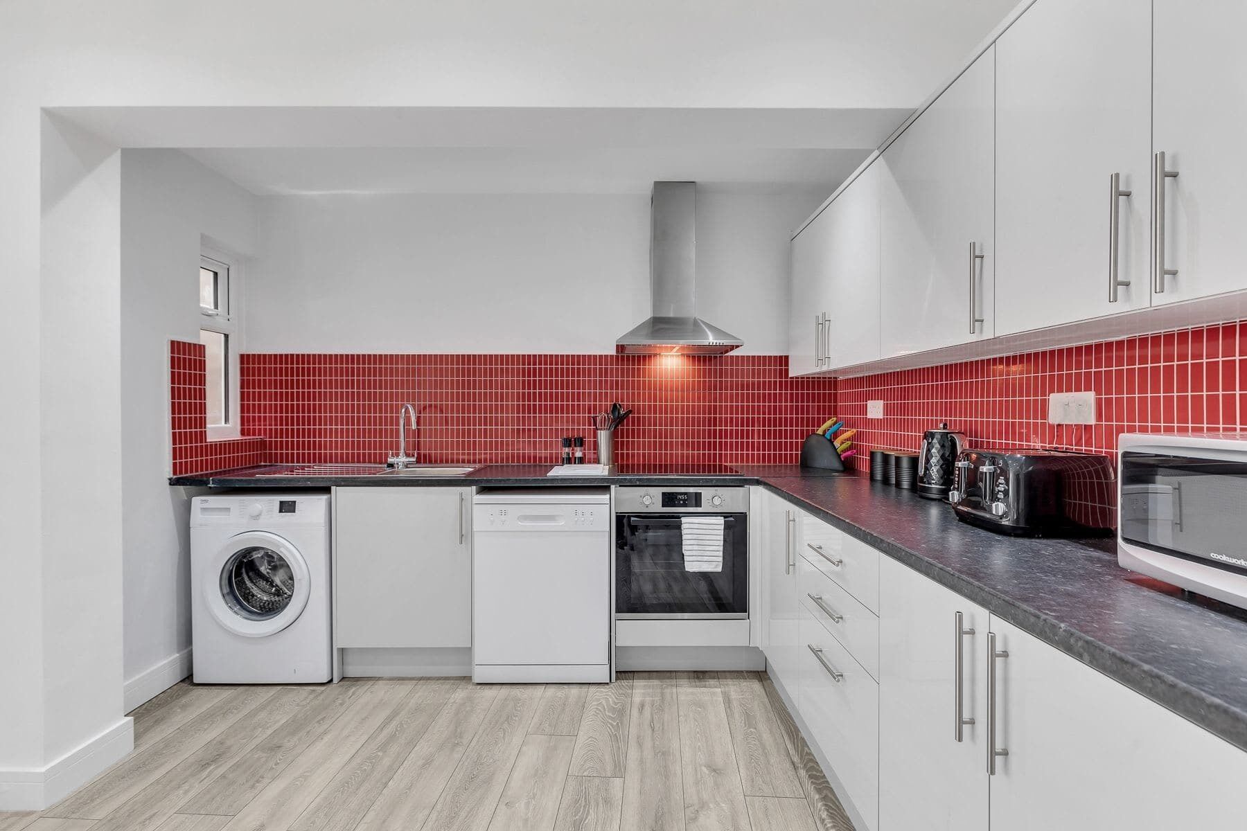 A compact kitchen with a washing machine, dishwasher, oven, and a red-tiled backsplash.
