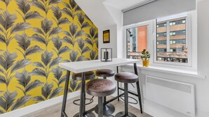 A modern kitchen with a white countertop, three stools, a window with a view of a building, and a yellow and gray leaf-patterned wall. - We Stay - Baxter Gate (Loughborough)