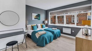 A modern bedroom with two beds, a desk, and a view of a brick building. - We Stay - Baxter Gate (Loughborough)