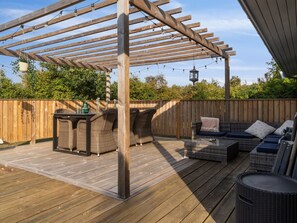 Relax on the deck with wicker furniture and a pergola overhead, surrounded by lush greenery.