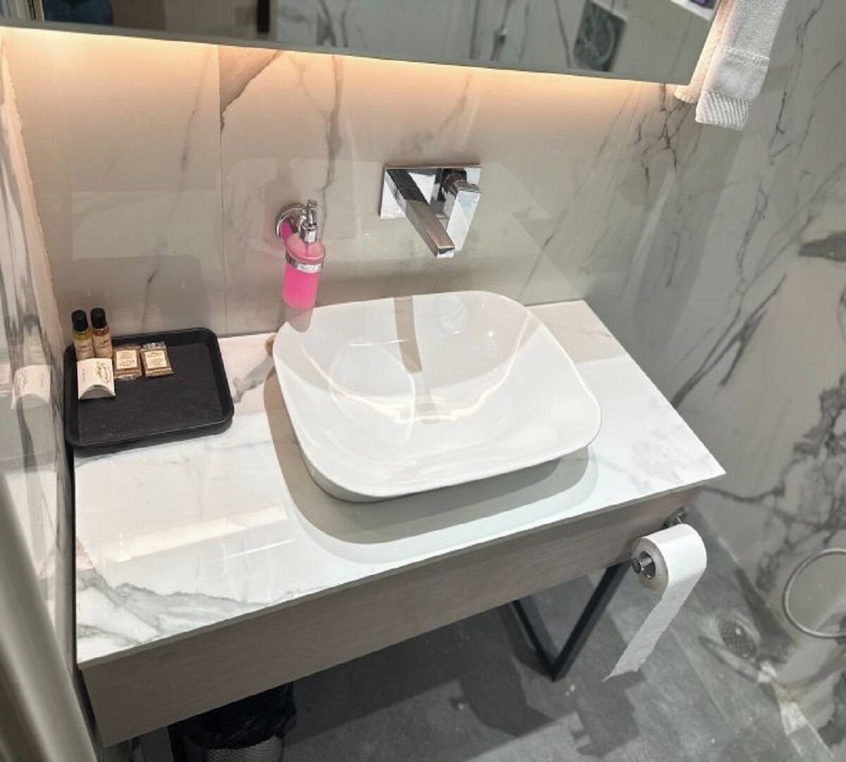 Un baño con lavamanos de mármol blanco, grifo cromado y bandeja negra con artículos de aseo.