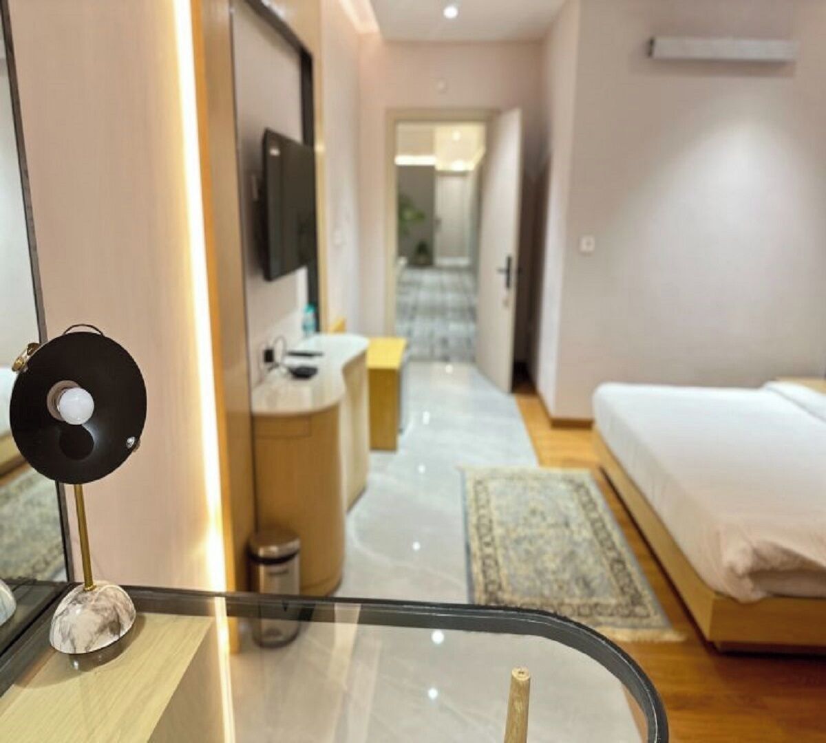 A modern hotel room with a glass table, a bed, a TV, and a view of the hallway.