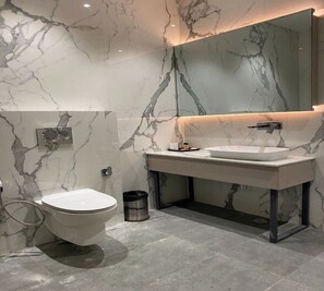 Un baño moderno con pared de mármol, inodoro blanco, lavabo con espejo y ducha.
