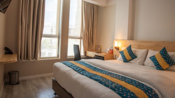 Ein Hotelzimmer mit einem großen Bett, einem Schreibtisch und einem Stuhl. Es gibt ein Fenster mit Vorhängen, einen Mülleimer und einen Nachttisch mit einer Lampe.