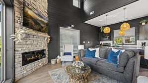 A modern living room with a stone fireplace, a grey sofa with blue cushions, a round coffee table, and a kitchen area with dark cabinetry and pendant lighting. - Luxe at Lake Haven - Lakefront pool & hot tub (Sevierville)