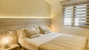 A bedroom with a wooden headboard, a bed with white linens and patterned pillows, a bedside table with a lamp, and a window with blinds. - Country House 'L Heura' with Private Terrace, Wi-Fi and Air Conditioning (Navata)