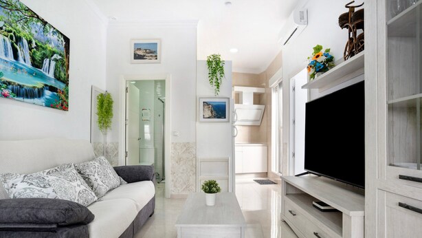 A modern living room with a grey sofa, a coffee table, a TV, and a kitchen area with white cabinets. - Holiday Home "Casa Centro Historico Barbate" with Private Terrace and Wi-Fi (Barbate)