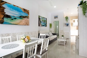 A dining area with a white table and chairs, a painting of a beach, a sofa, and a small table with a plant.