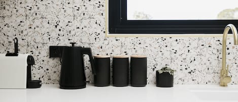 A kitchen with a white countertop, a black coffee maker, three black containers, a small potted plant, and a gold faucet.