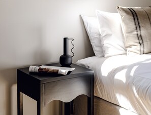 A neatly made bed with white linens and a striped pillow, a black bedside table with a vase and a magazine, and a plain wall in the background.