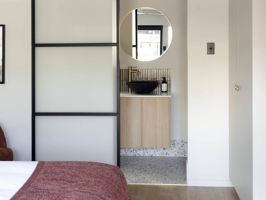 A modern hotel room with a bed, a brown armchair, a framed abstract painting, a bathroom with a sink and mirror, and a door leading to another room.