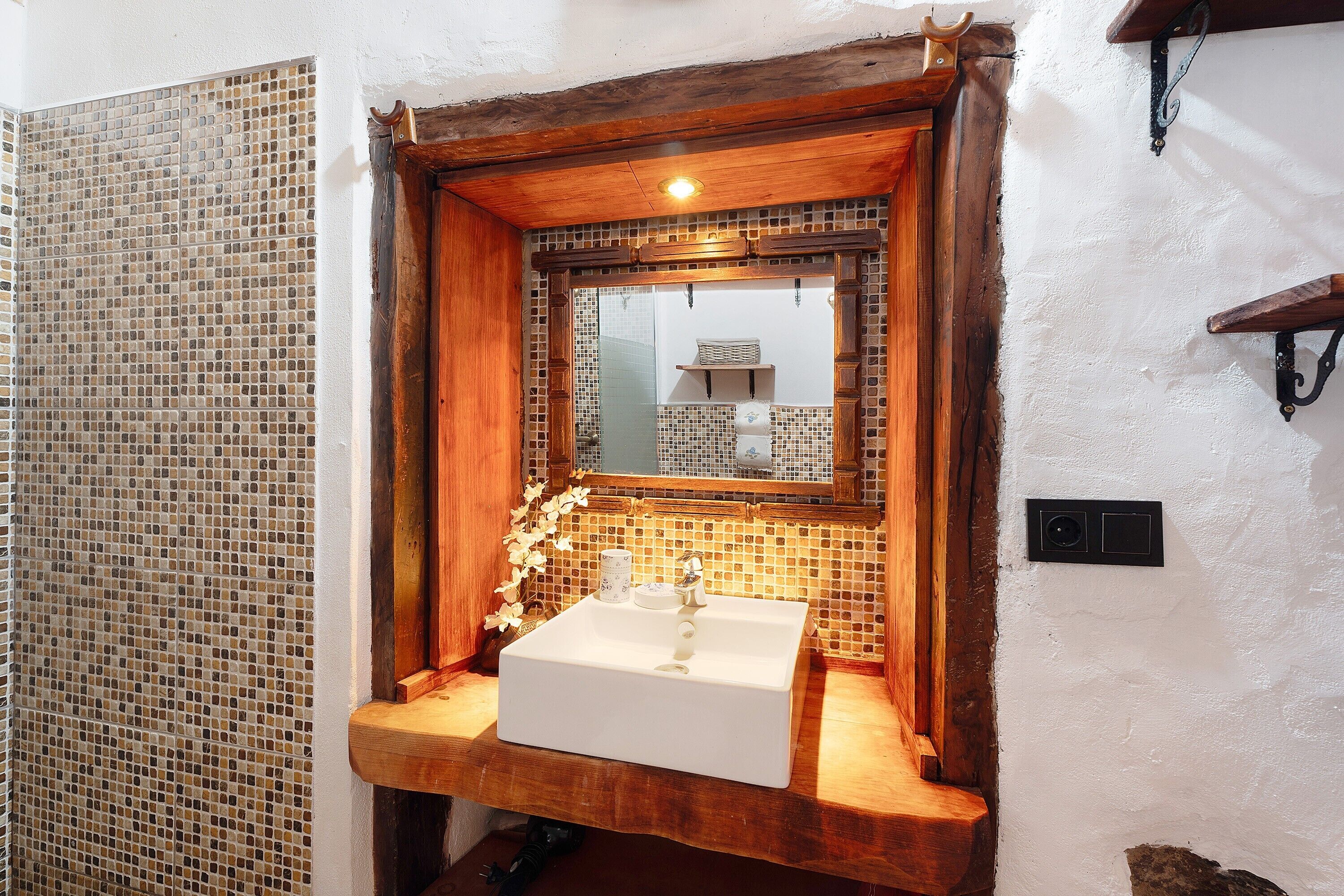 Un baño con lavamanos blanco, tocador de madera, salpicadero de mosaico y espejo con marco de madera.