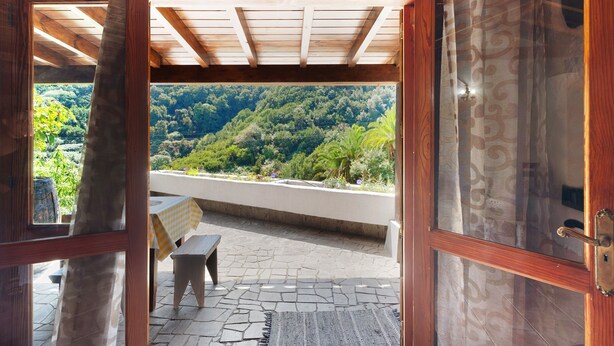 Eine offene Holztür führt zu einem Balkon mit Steinboden, einer Bank und einem Tisch mit kariertem Tischlein.