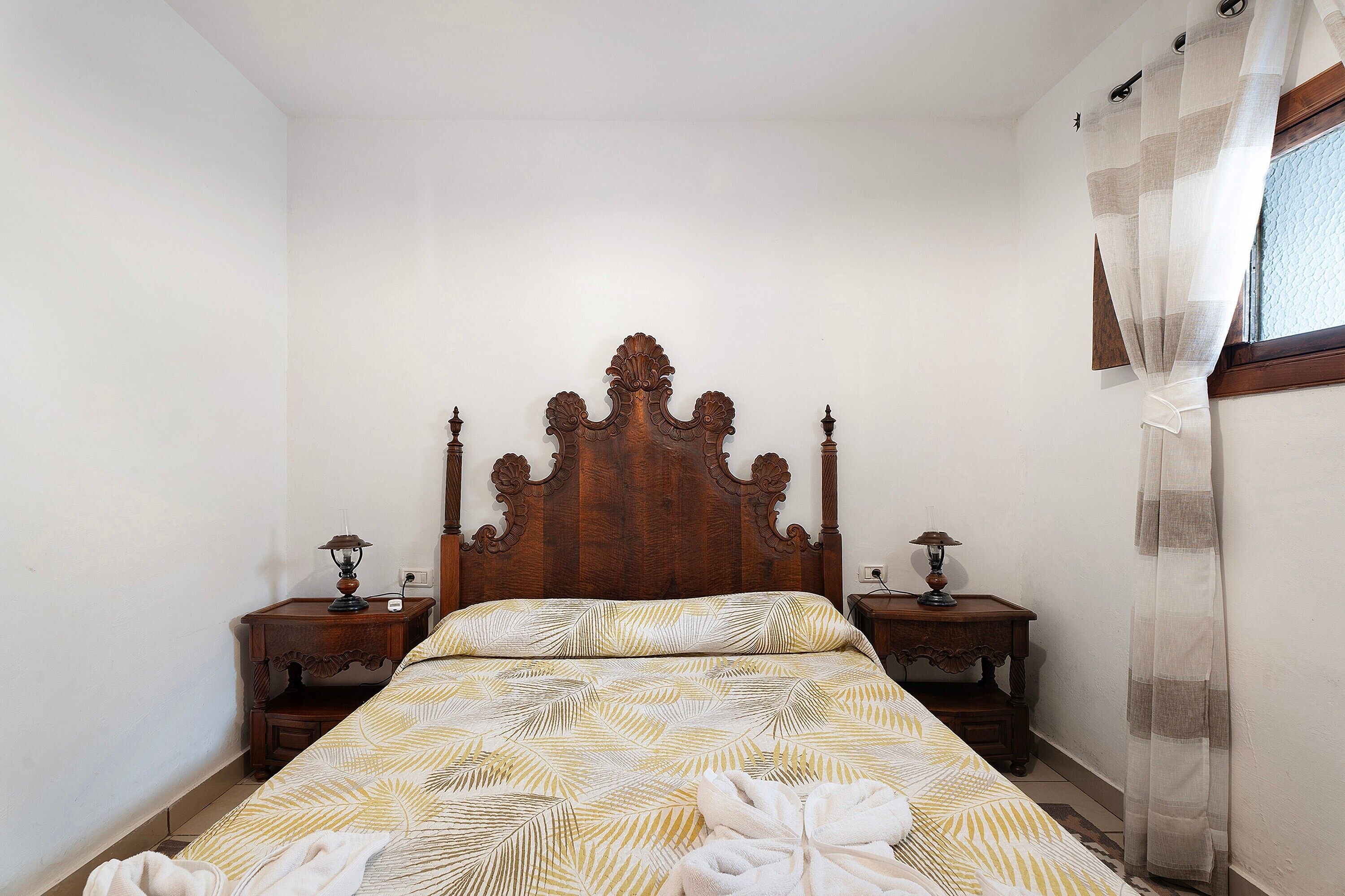 A bedroom with a wooden headboard, two bedside tables with lamps, a bed with patterned bedding, and a window with sheer curtains.