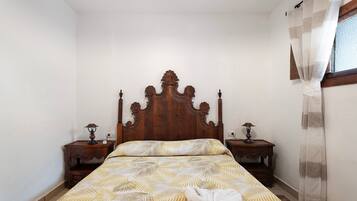 A bedroom with a wooden headboard, two bedside tables with lamps, a bed with patterned bedding, and a window with sheer curtains.