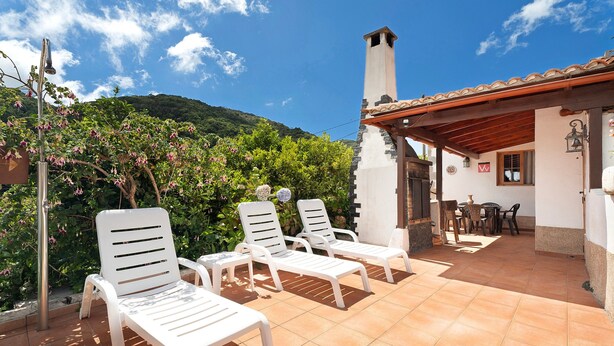 A patio with white lounge chairs, a tiled floor, a small table, and a building with a chimney.