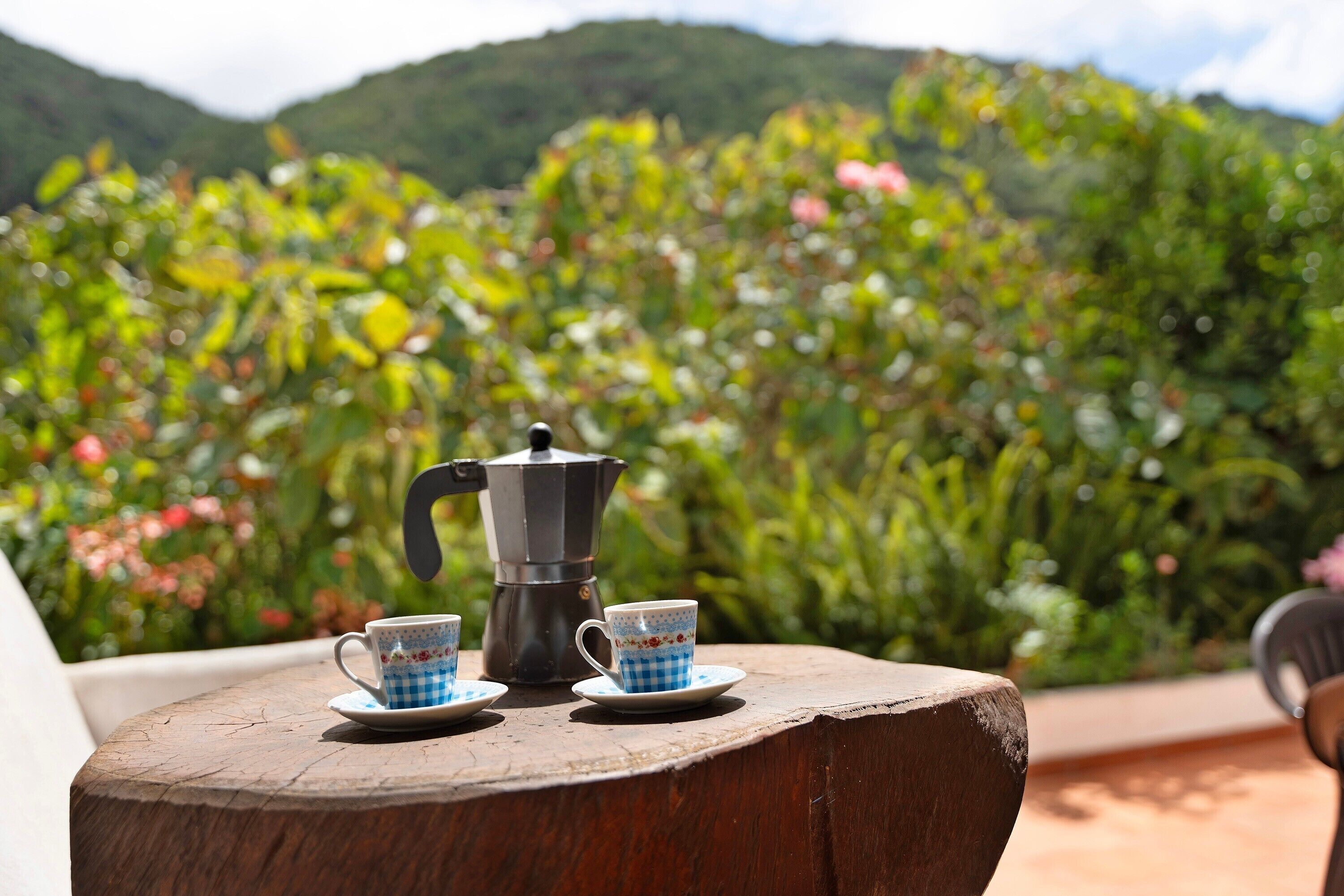 A rustic wooden table with a coffee maker and two cups, set against a backdrop of lush greenery and hills.