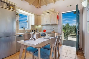 A kitchen with a dining table, chairs, a refrigerator, and a window with a view of trees. - Villa 'Belvedere Rosa' with Sea View, Pool, Terrace & Wi-Fi (Lefkada)