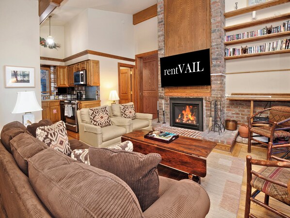 A spacious living room with a fireplace, a kitchen in the background, and a bookshelf filled with books.