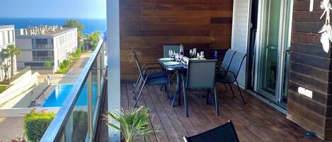 A balcony with a wooden floor, a table with chairs, a potted plant, and a view of the ocean and buildings.
