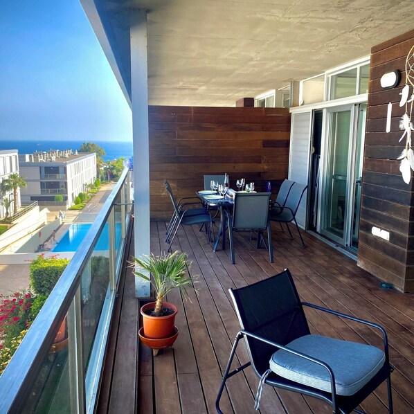 A balcony with a wooden floor, a table with chairs, a potted plant, and a view of the ocean and buildings.