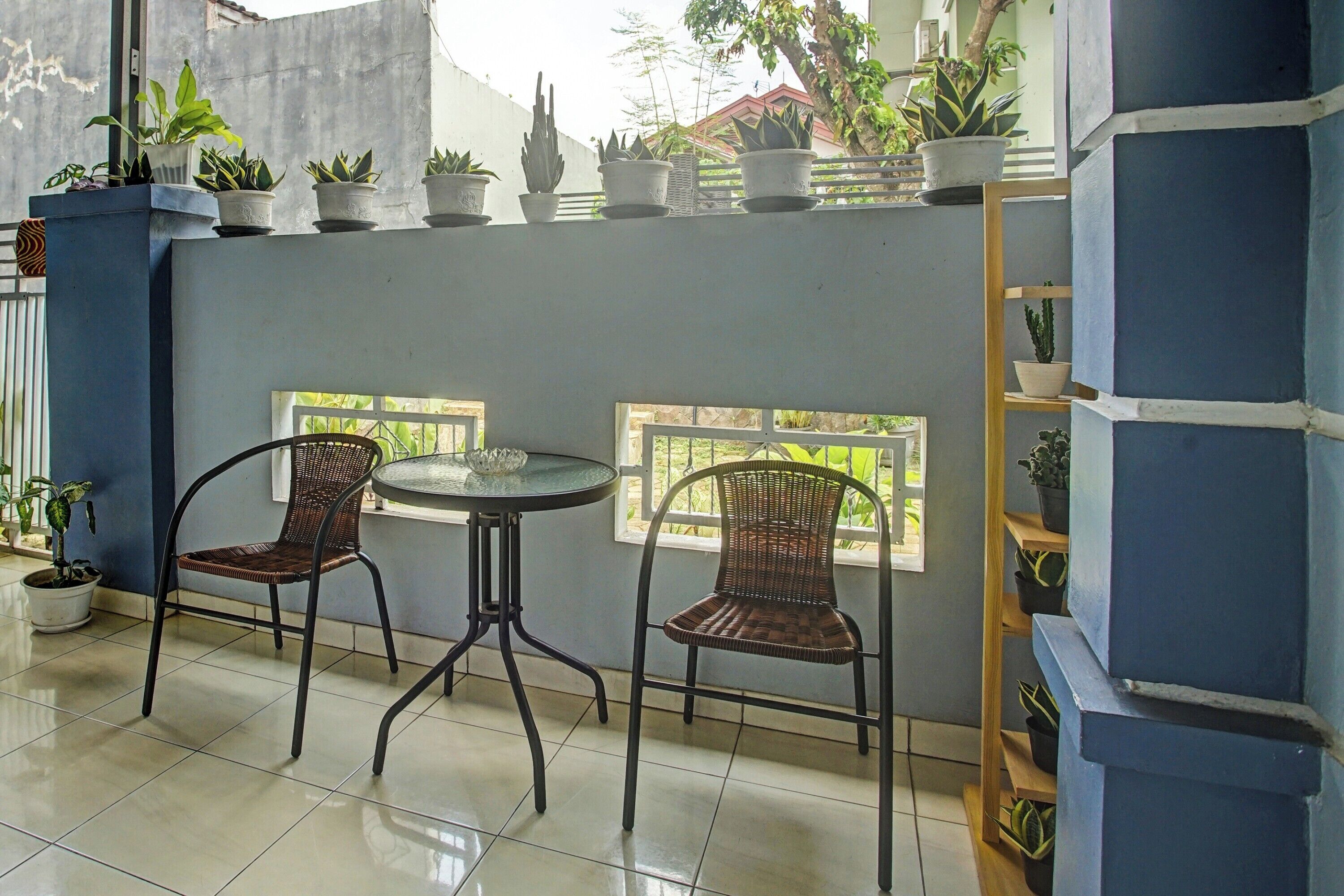 A small, enclosed patio area with two wicker chairs around a glass table, surrounded by potted plants and a view of greenery outside.