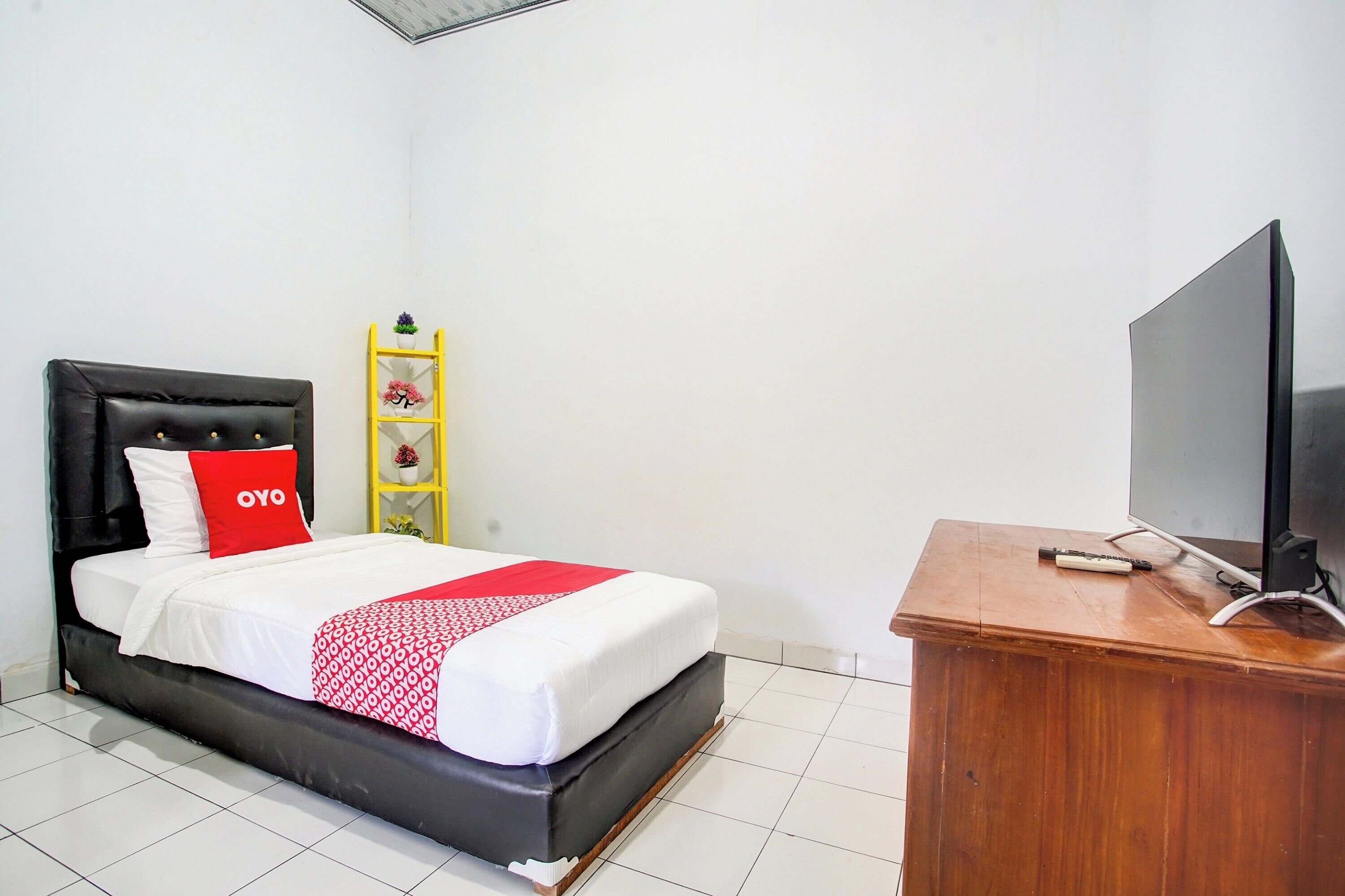 A neatly made bed with a red pillow, a wooden dresser with a television, and a yellow shelf with decorative items.