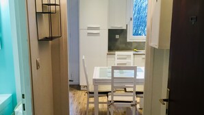 Een smalle gang die uitkomt in een keuken met witte kasten, een eettafel en stoelen.
