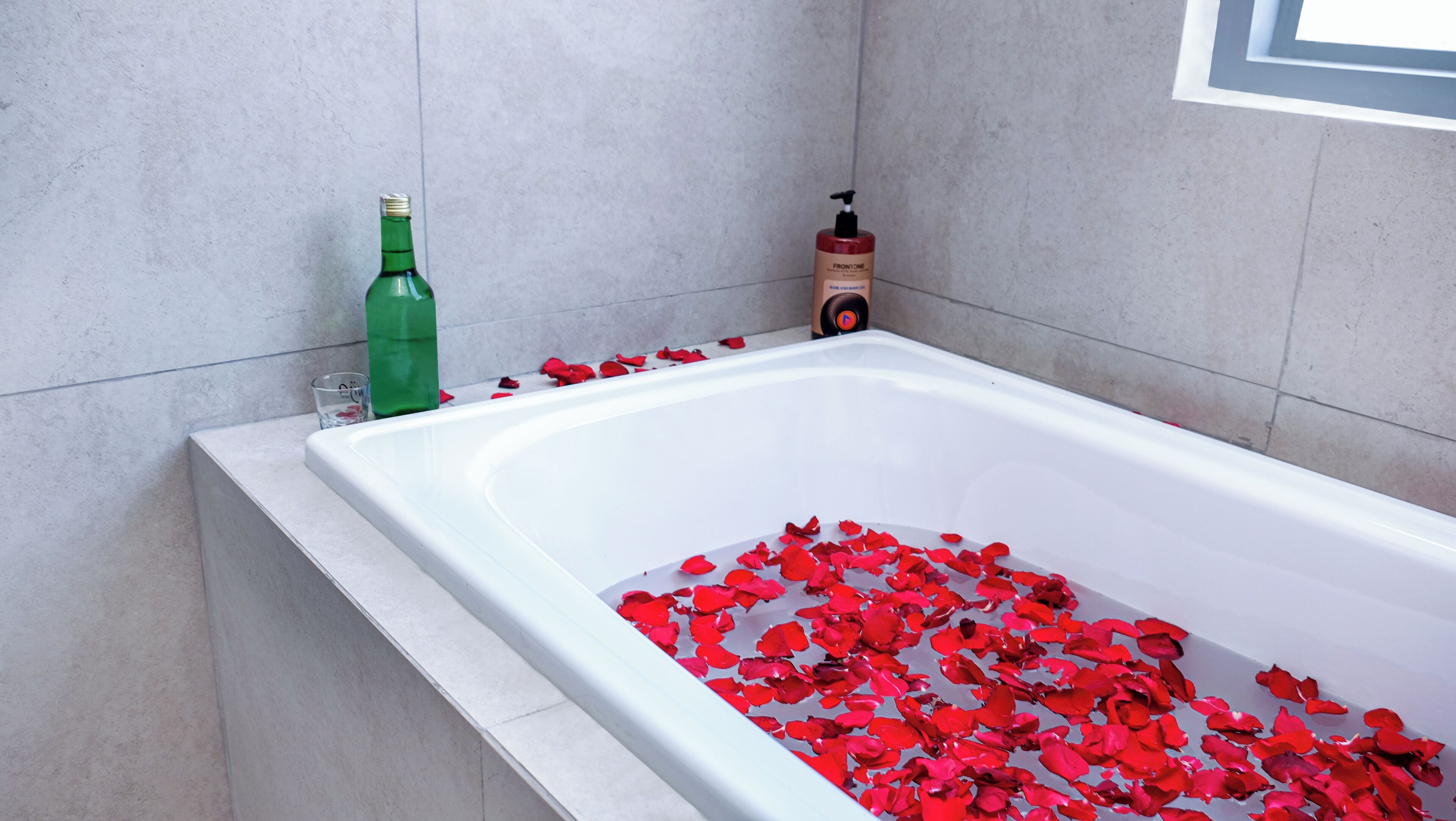 A bathtub filled with red rose petals, a green bottle, and a red bottle on the side.
