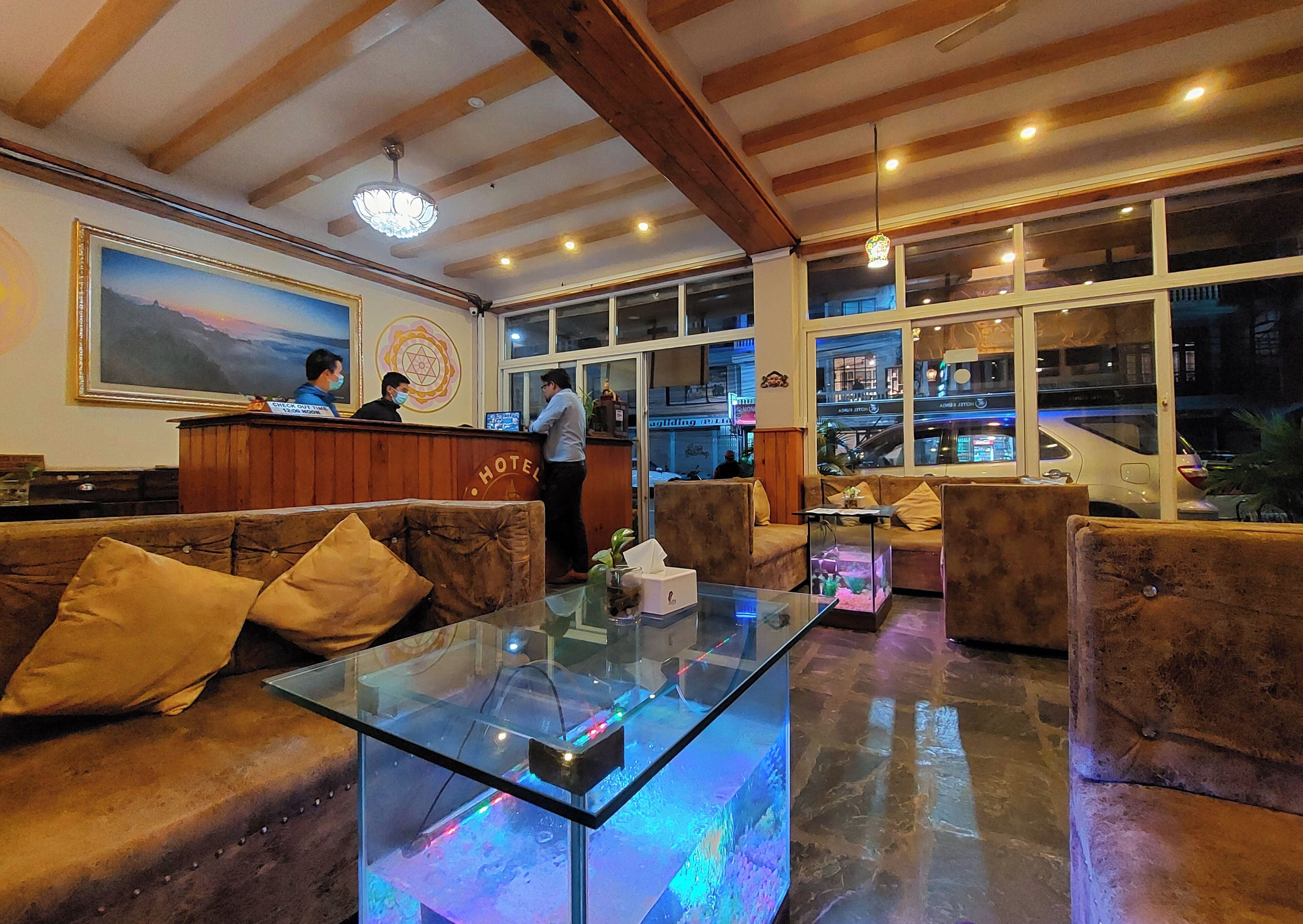A hotel lobby with wooden beams, a reception desk, a glass coffee table, and a large window with a view of the street.